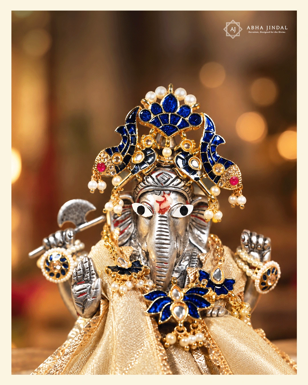 Front view of Ganesh Ji in a patterned gold-tone poshak with a deep blue lotus and fish-motif mukut, coordinated blue adornments and pale bead-like drops.