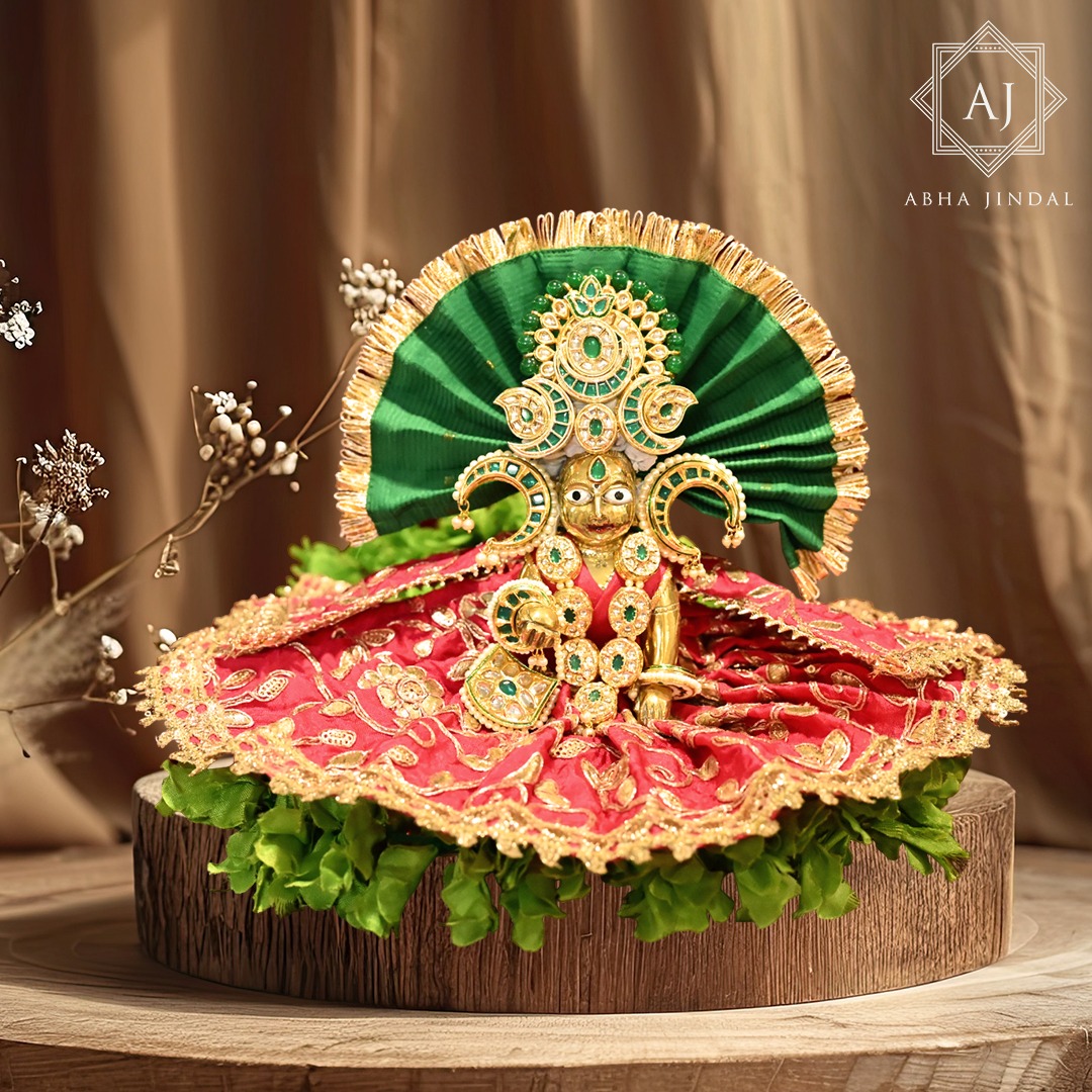 Thakurji in red embroidered poshak with a green fan-style presentation, green and clear mukut details and gold-tone adornments on a wood display base