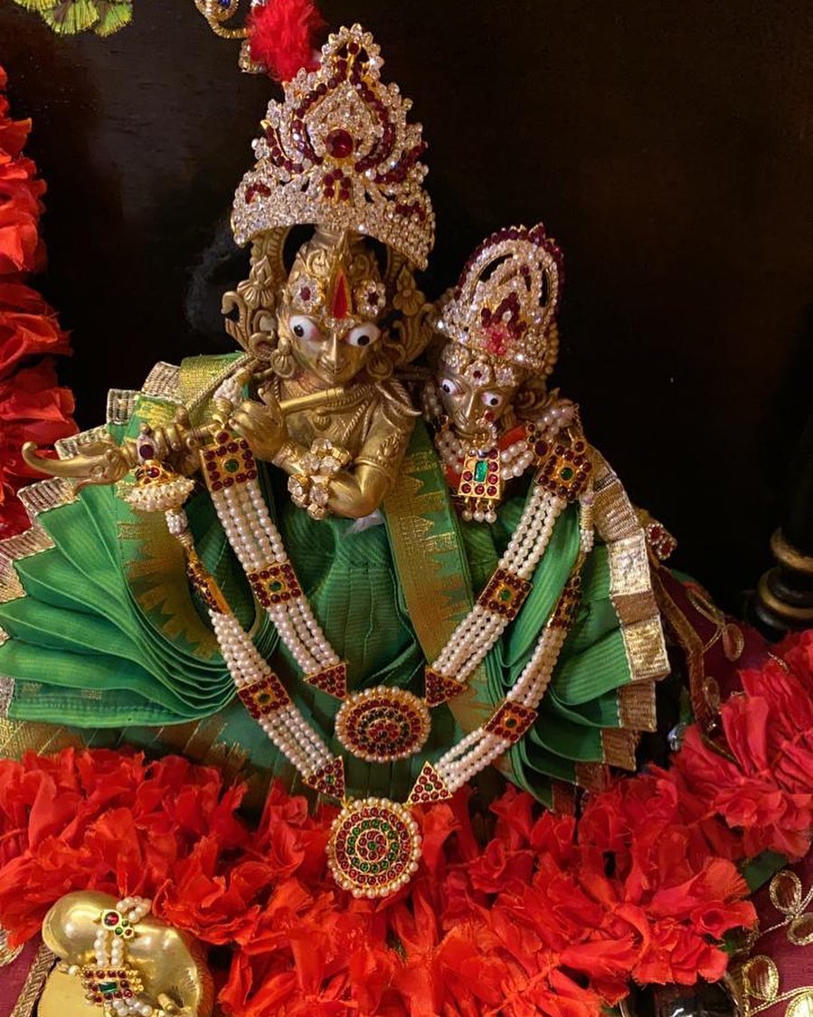 Radha and Krishna pair in green poshaks with ornate crowns and long white-beaded necklaces above red flowers