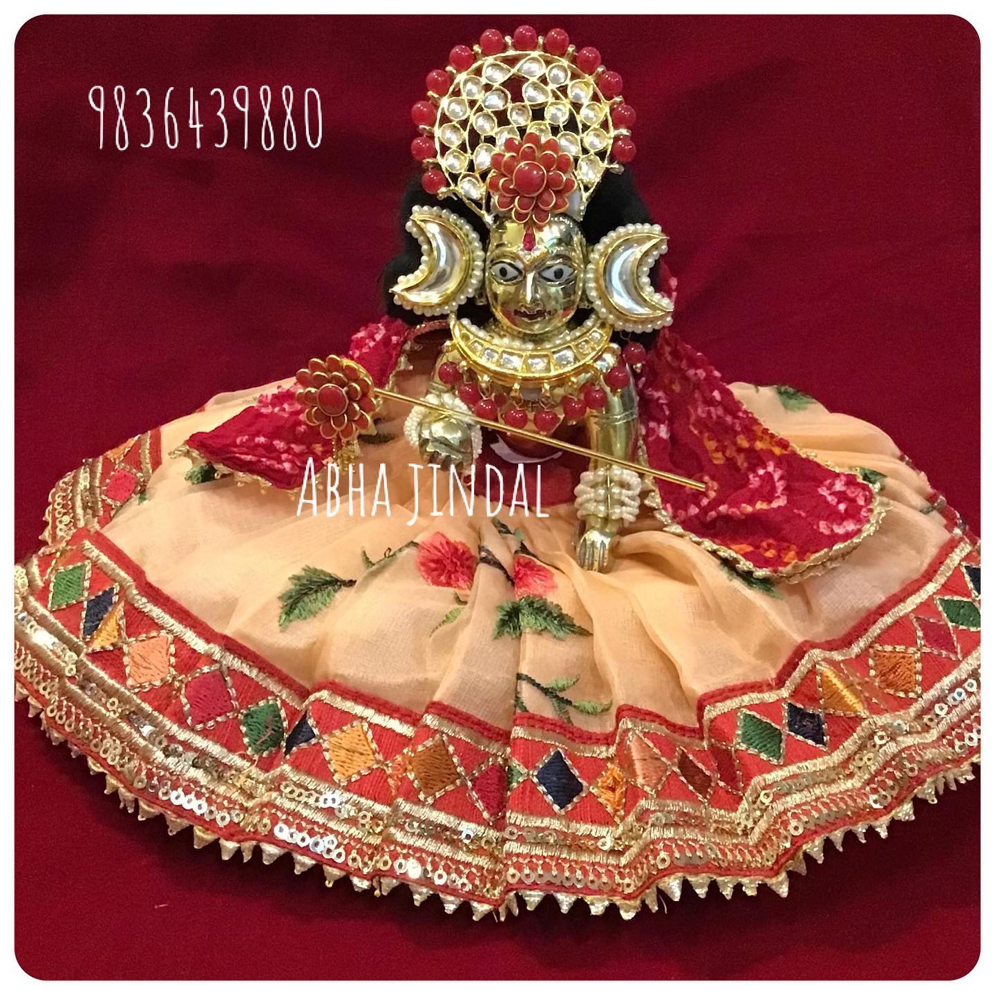 Gold-tone deity in a beige poshak with maroon geometric border, red drape and circular head adornment