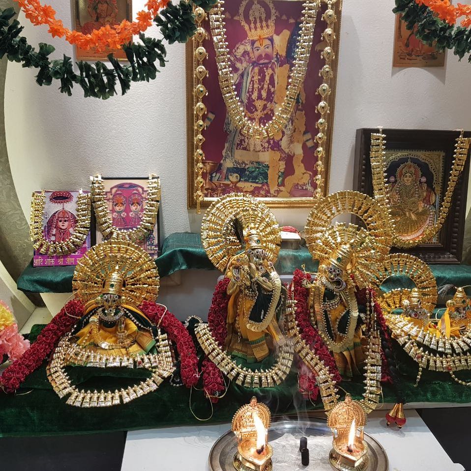 Several deity figures in fan-style poshaks and gold-tone head adornments before a home altar