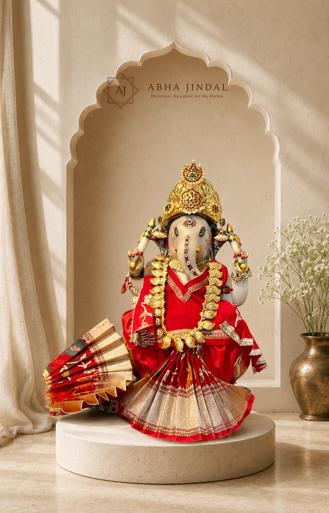 Seated Ganesh Ji in red and white attire with floral garlands and a fan-pleated base