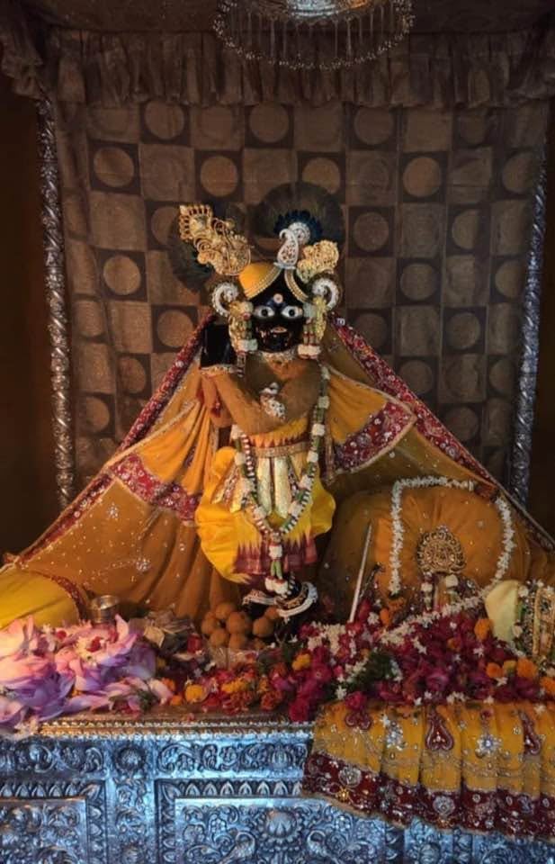 Front shrine view of Banke Bihari in orange and gold-toned attire with a flute and floral offerings.