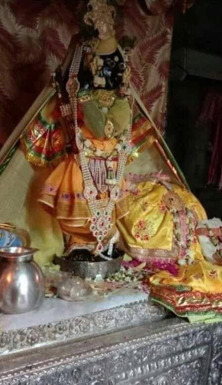Low-light sanctum photograph of Banke Bihari in yellow, orange and green attire with layered jewellery.