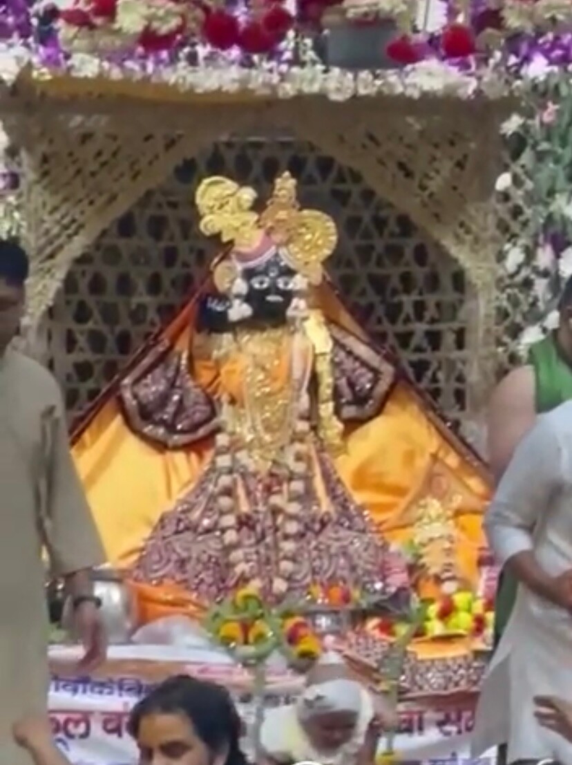 Banke Bihari shrine with deity in yellow and maroon attire, gold-toned ornaments, flowers, attendants and visitors.
