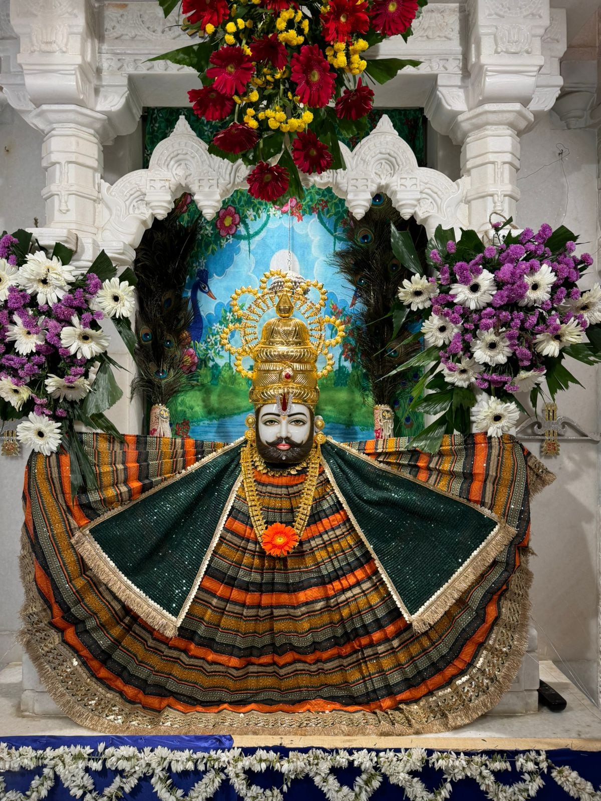 Shyam Baba shrine figure in a black and orange fan-shaped poshak inside a white marble niche.