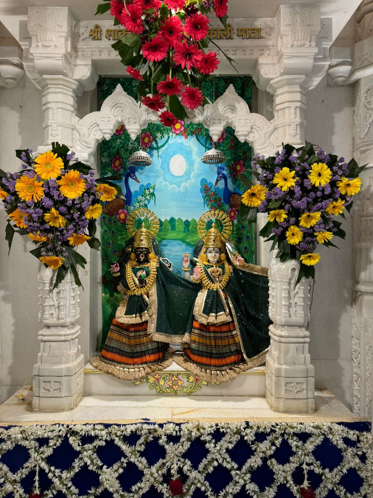 Two crowned shrine figures in coordinated black and orange poshak inside a white marble niche.