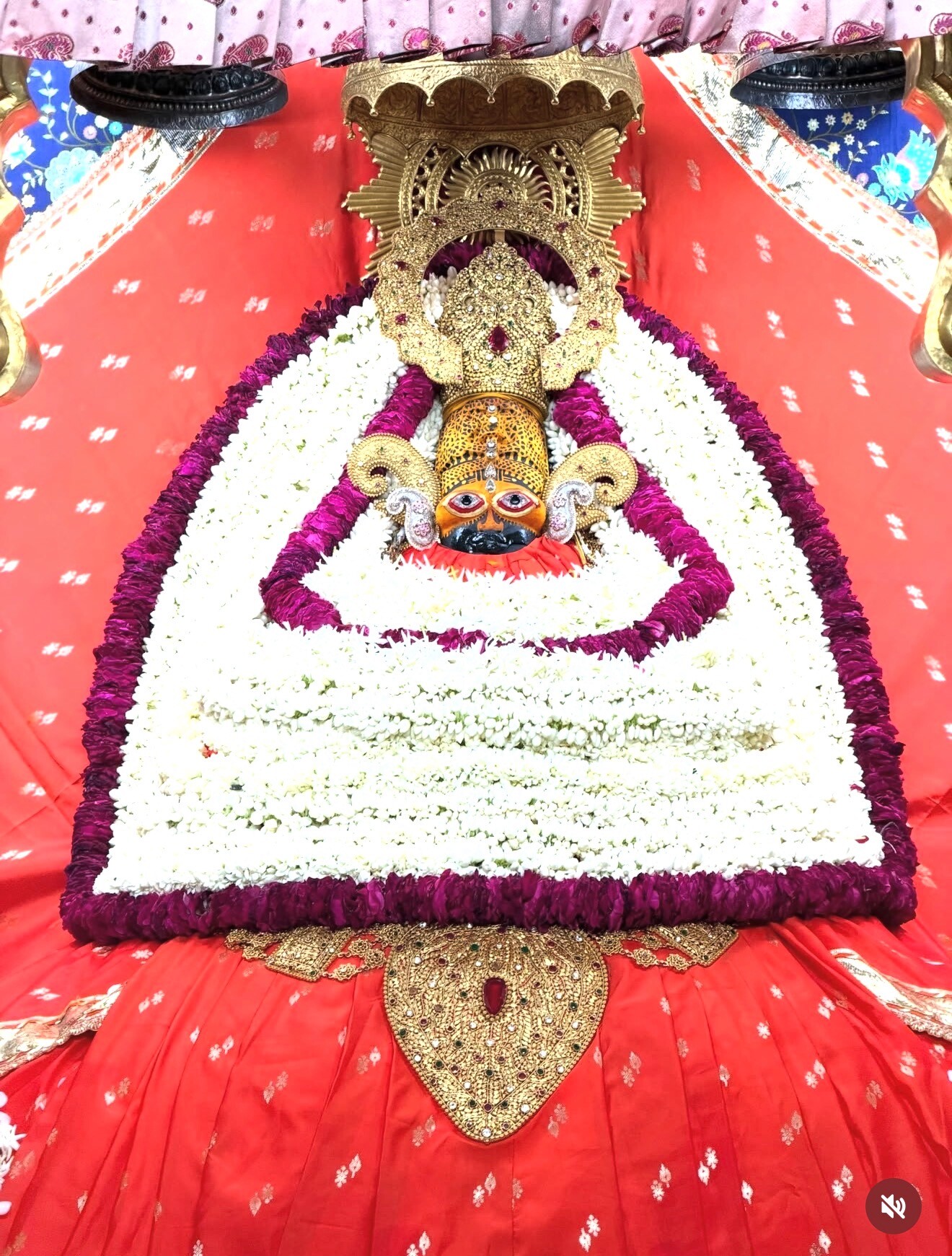 Close view of Shyam Baba in red poshak with gold border, white and magenta flowers, gold-toned adornments and a gilded shrine.