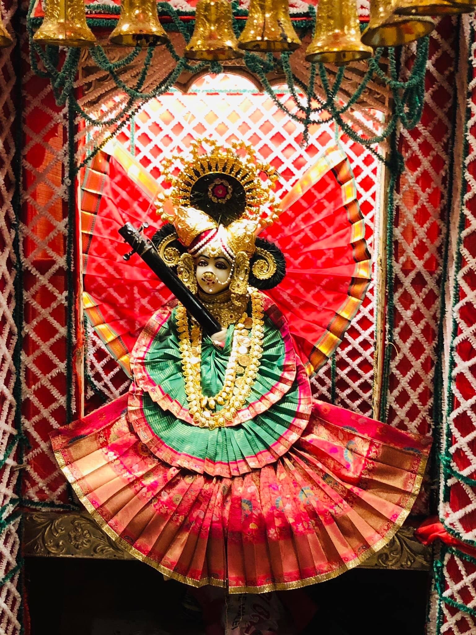 Nidhivan shrine figure in green and rose pleated attire beneath brass bells and garlands.